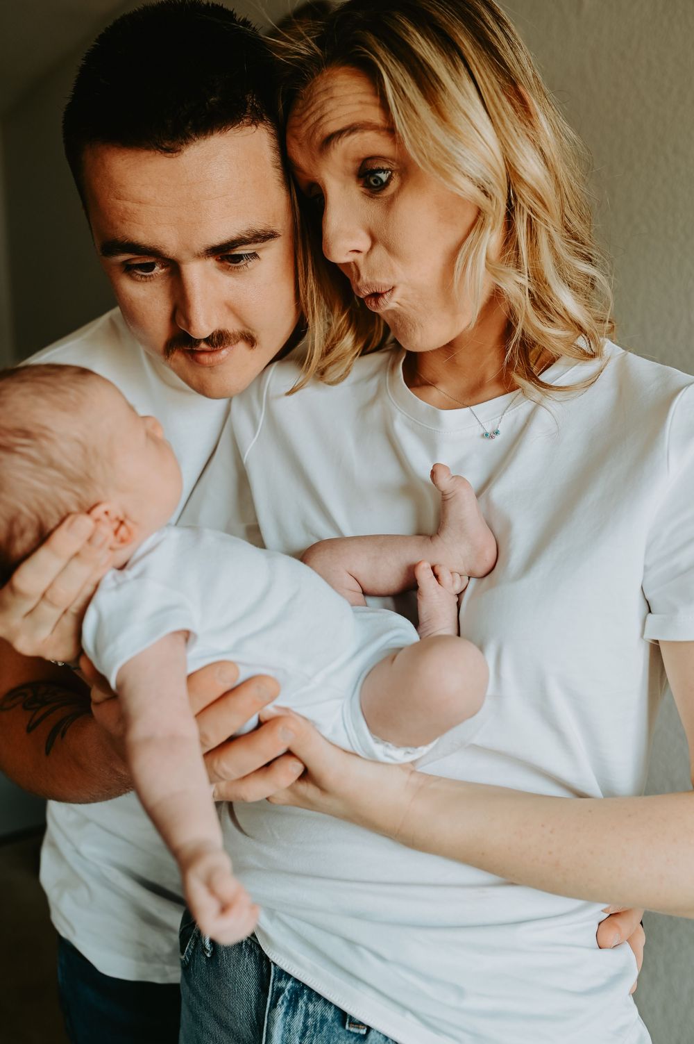 Young couple in white t-shirts lovingly holding their newborn baby, making funny faces together.