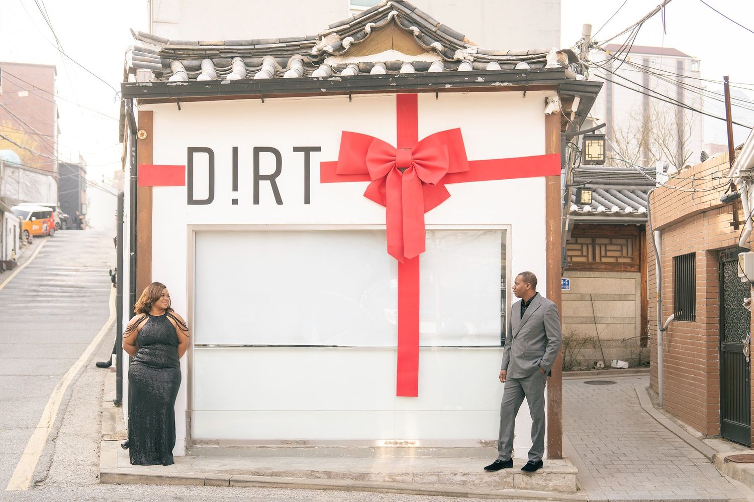Couple poses outside a white building with a giant red bow and DIRT sign in a Korean alleyway.