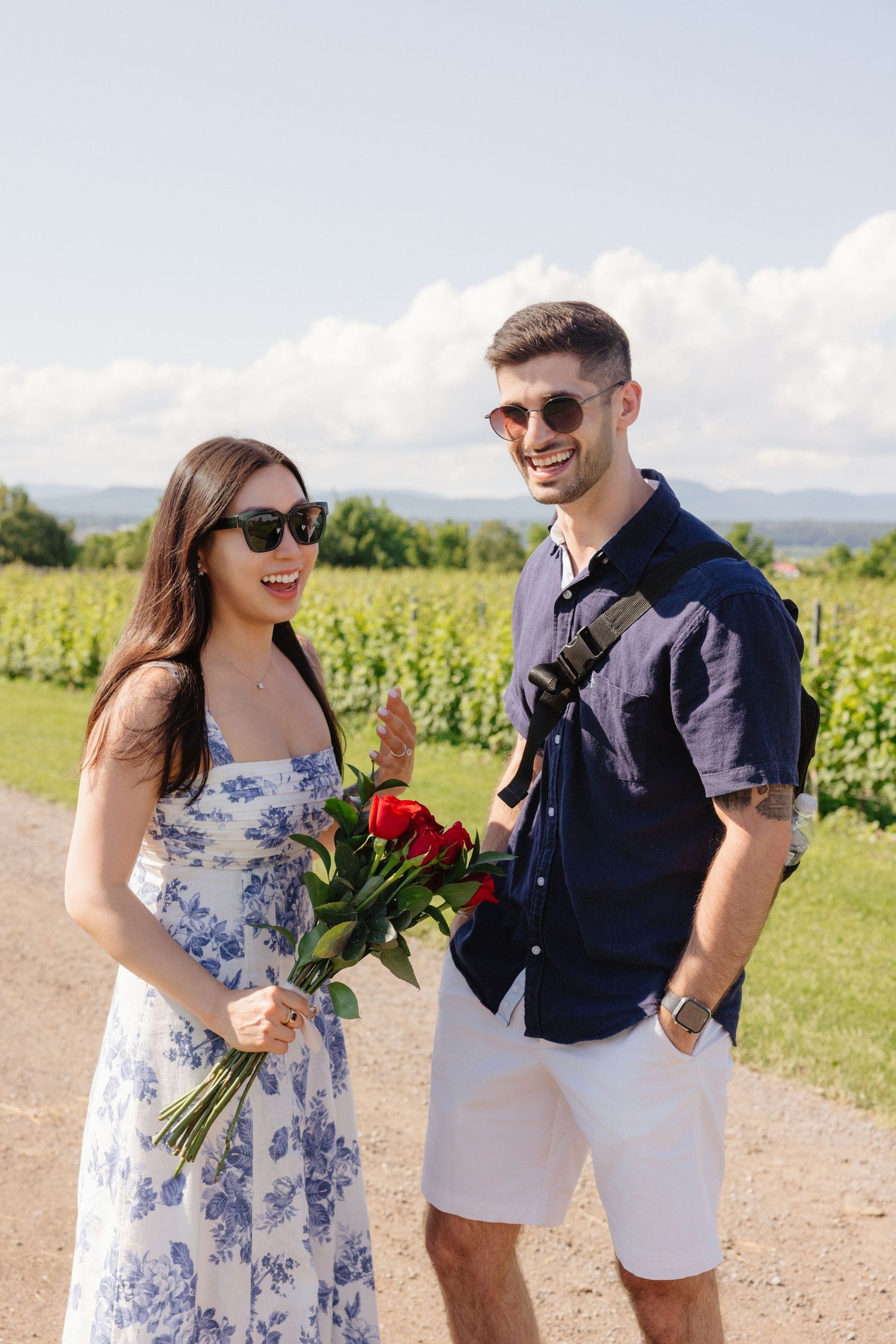 Couple in summer attire holding red roses in vineyard with mountains in background.
