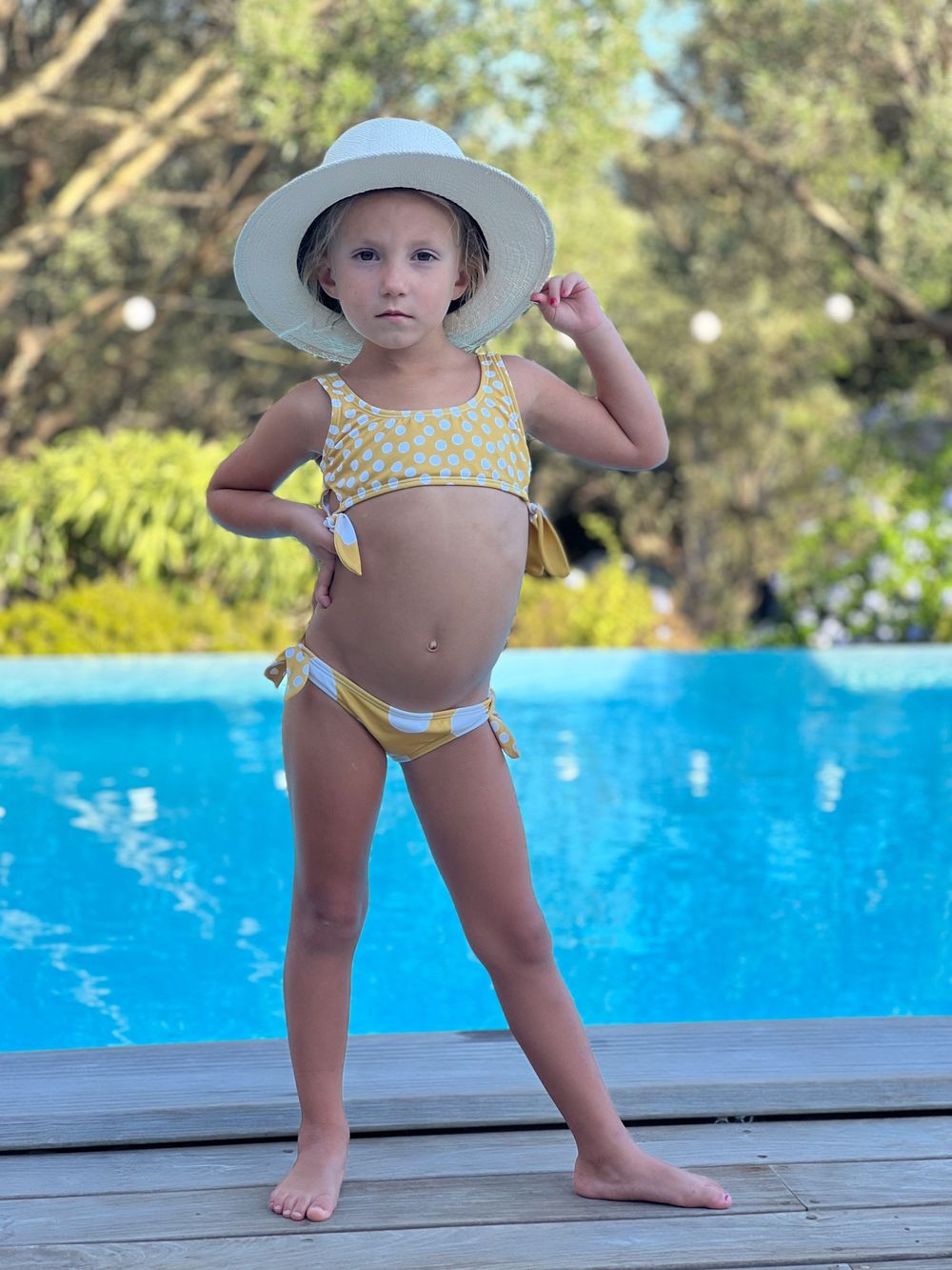Young girl in yellow polka dot bikini and white sun hat posing by a blue swimming pool on a wooden deck.