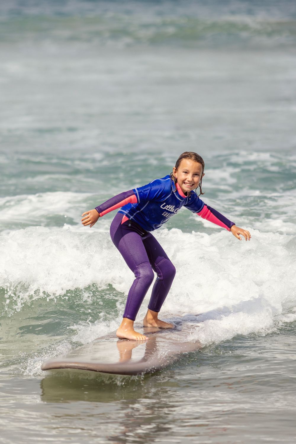 Young girl in purple wetsuit smiling while surfing a wave on a longboard at the beach.