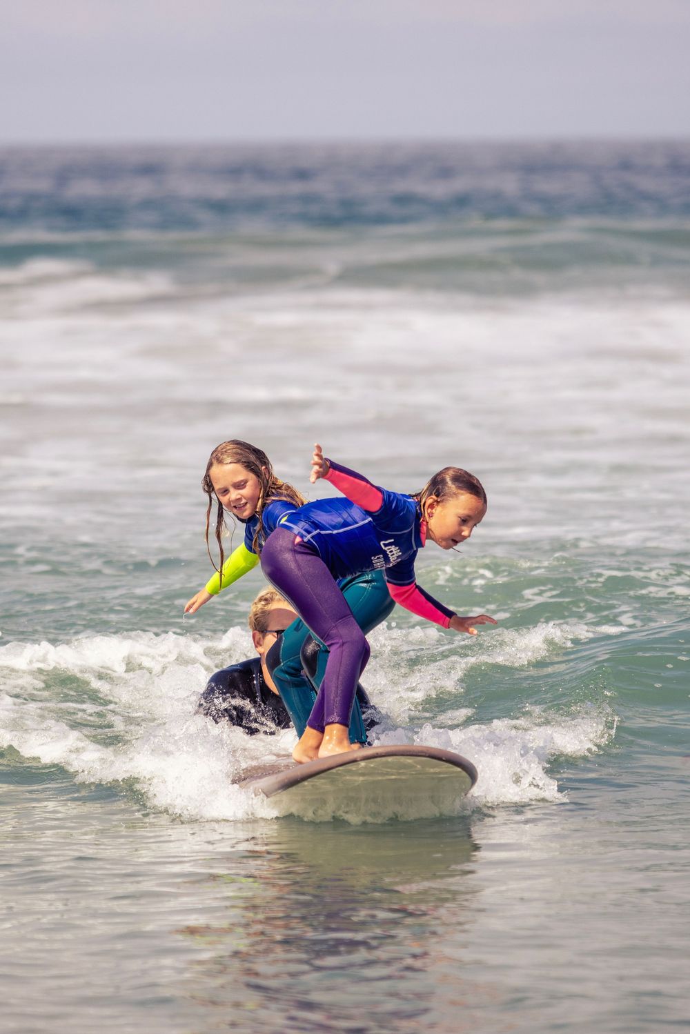 Two young girls in wetsuits laugh while riding a surfboard together on ocean waves.