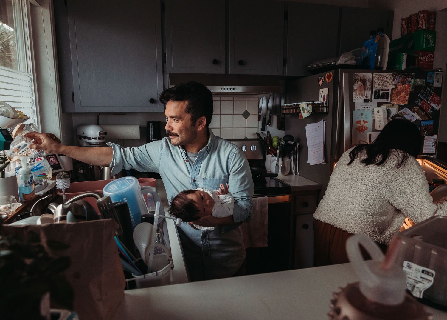 Man multitasking in kitchen, holding newborn baby while reaching for items at the sink.