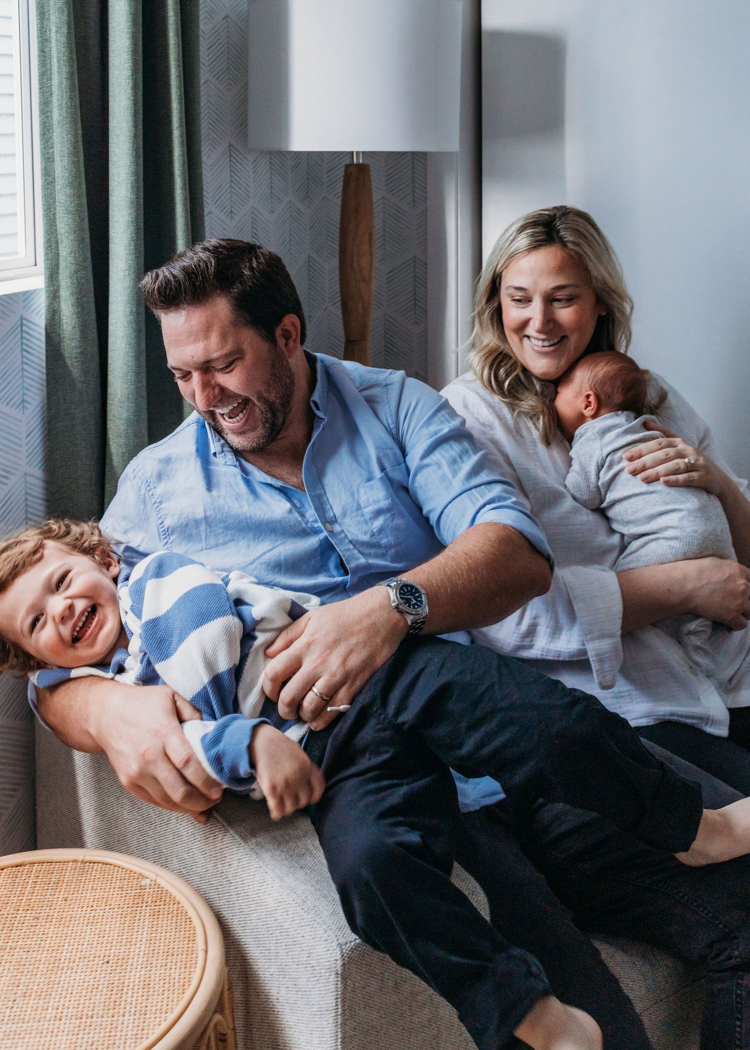 Happy family of four laughing together on a couch, parents holding children in a bright cozy living room.