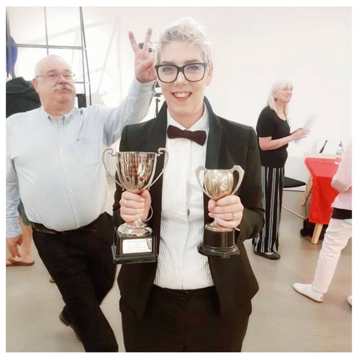 Young person in tuxedo holding two silver trophy cups, smiling and making a victory sign at an awards event.