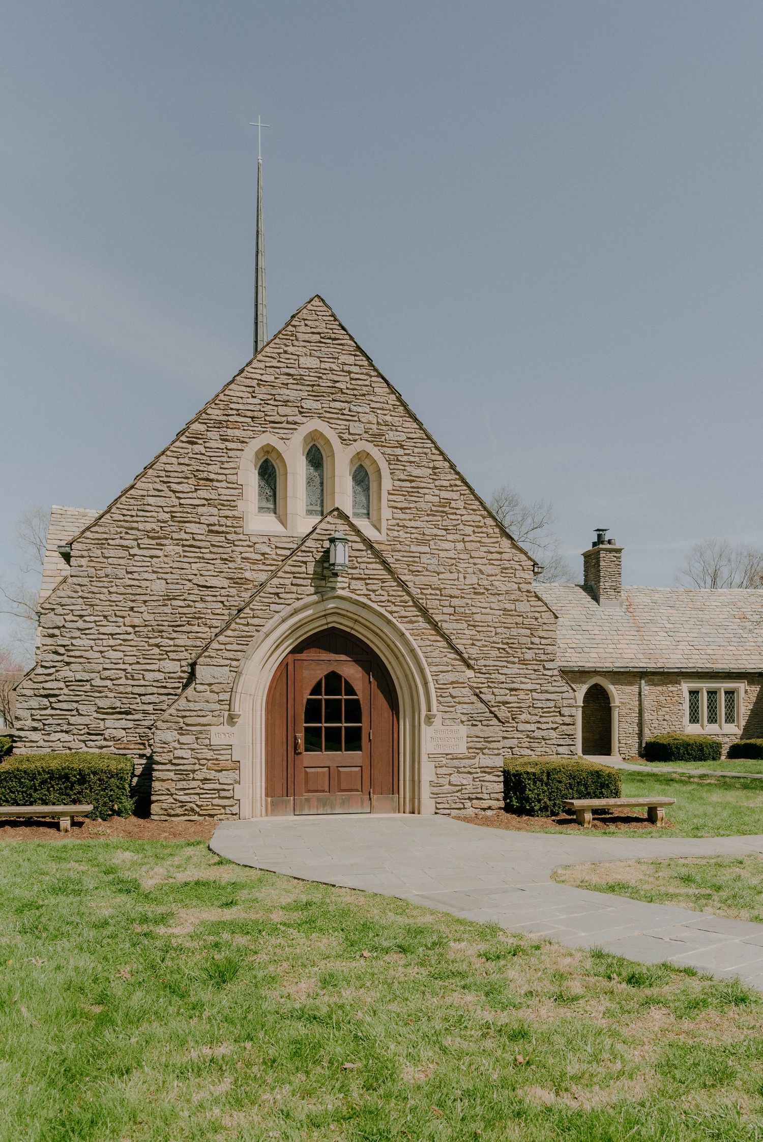 Stone church with Gothic arched entrance, steeple, and manicured lawn surrounded by bare trees on a cloudy day.