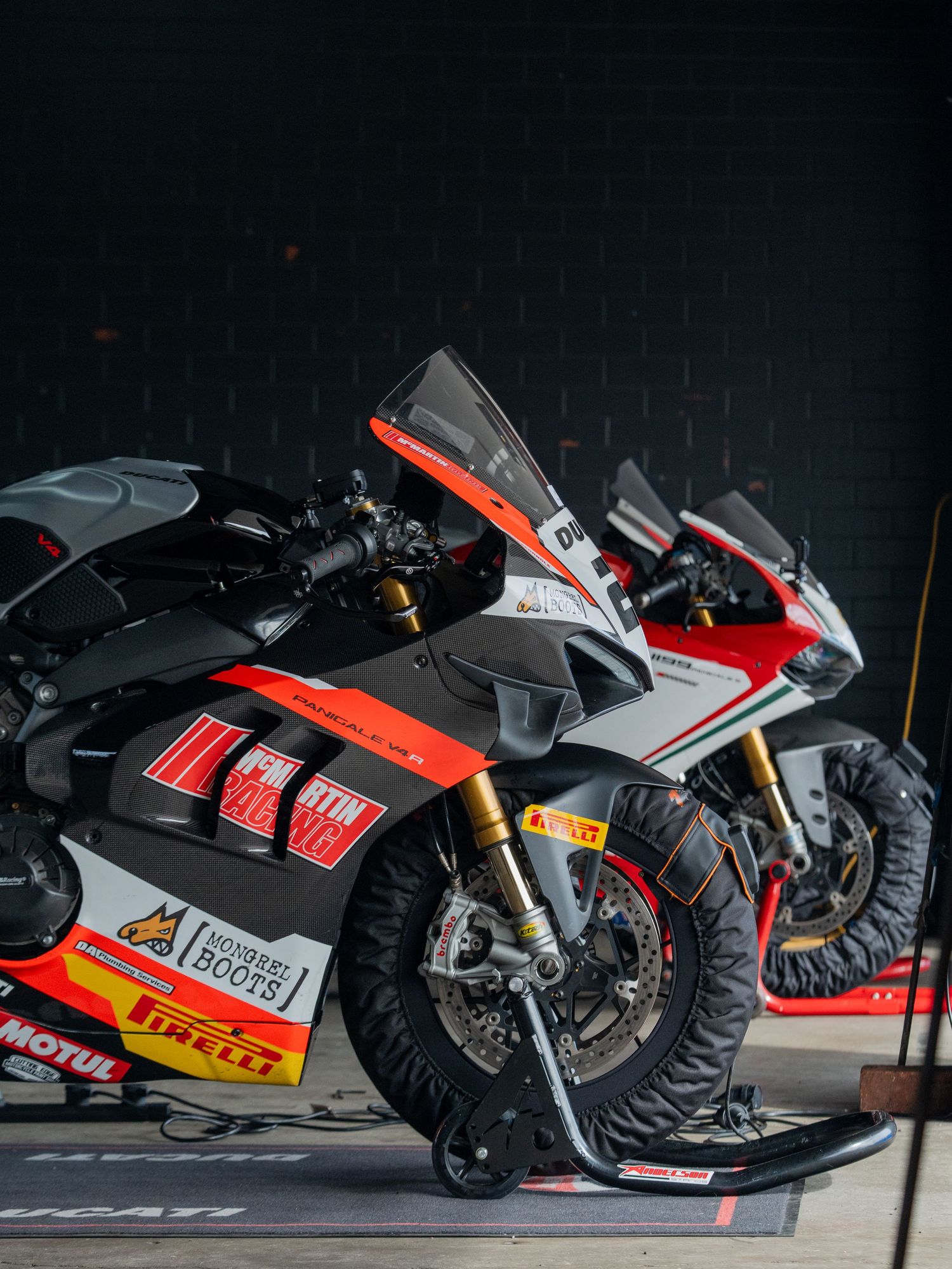 Red white and black Aprilia RSV4 superbike on tire warmers in a dark racing paddock garage.