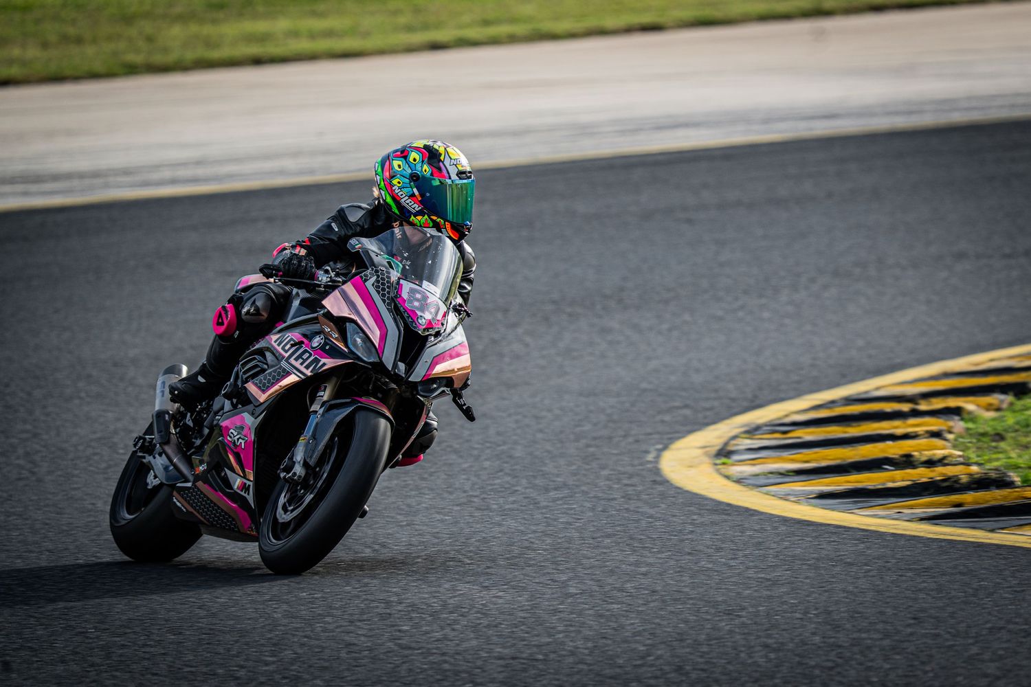 Motorcycle racer on pink and black BMW sportbike cornering aggressively on a race track.