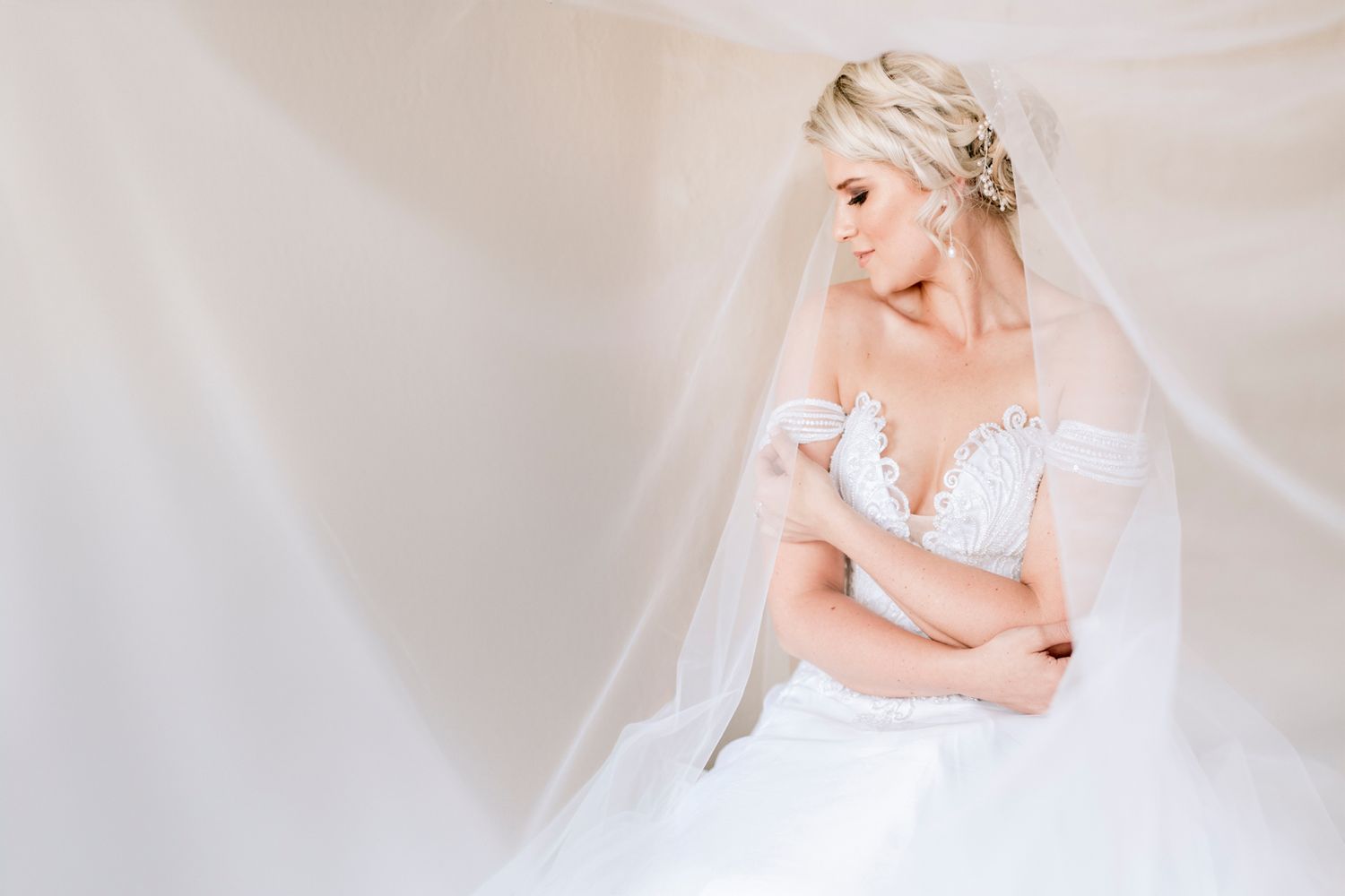 Elegant bride in off-shoulder lace wedding gown wrapped in flowing veil in soft natural light.