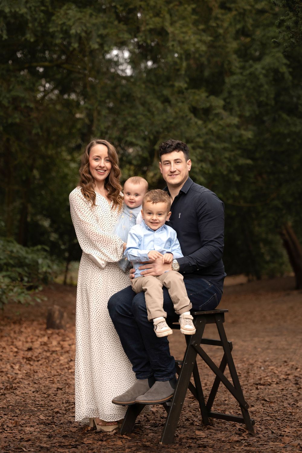 Family portrait of parents with two young boys in a woodland setting, mother in white dress, father in navy shirt.