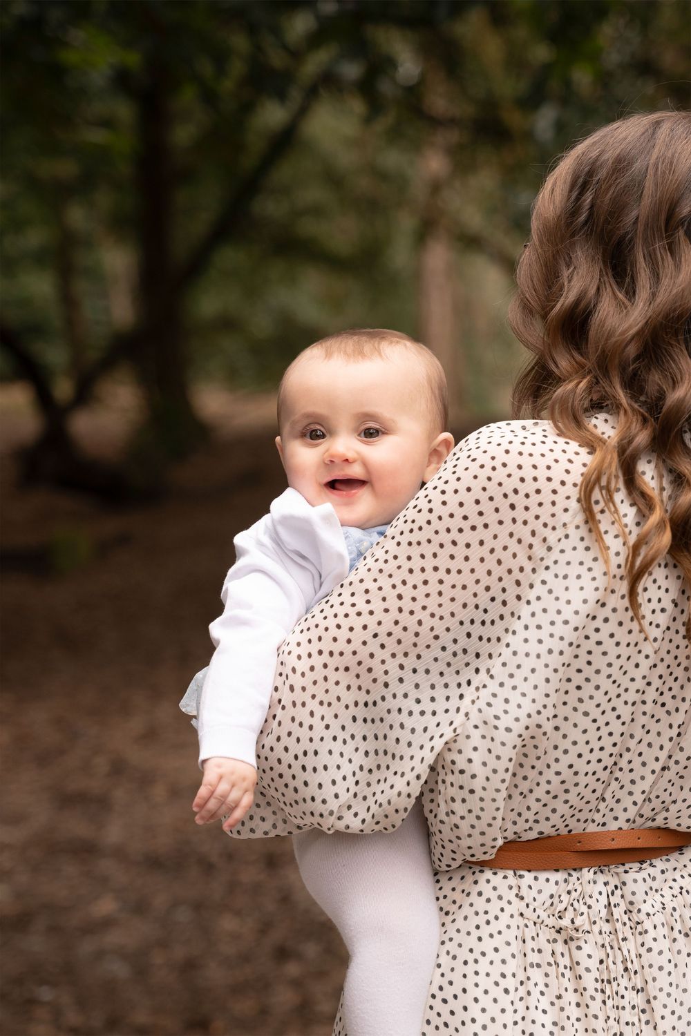 A smiling baby being held by a mother in a polka dot dress while walking through a forest path.