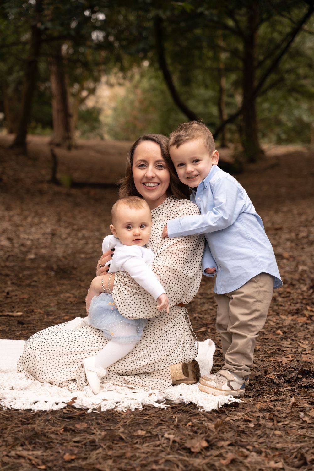 Mother sitting on blanket in woodland, smiling with baby and toddler son hugging her from behind.