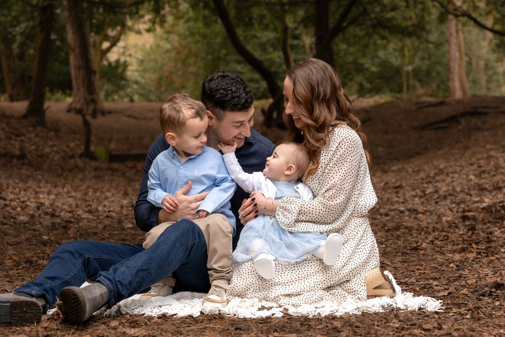 Family of four sitting on blanket in forest, parents bonding with toddler and baby in warm autumn setting.