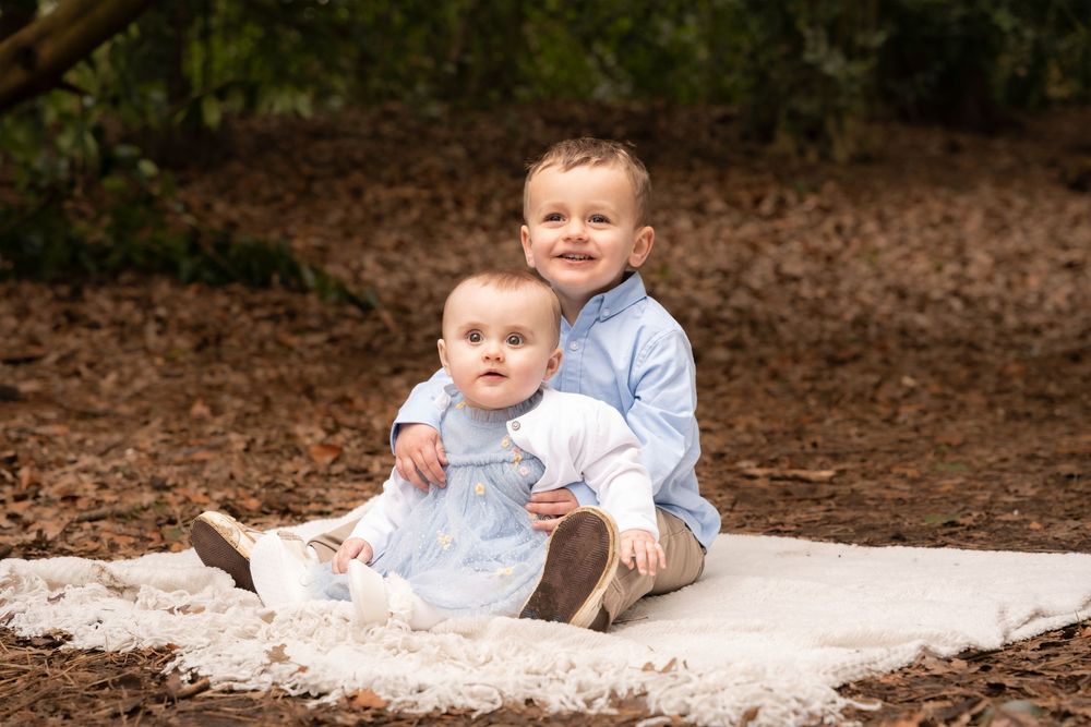 Two young siblings sit on a white blanket outdoors, older toddler in blue hugging baby in white dress.