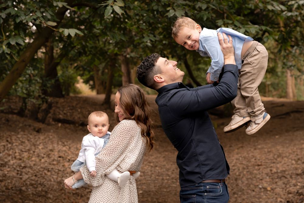 Joyful family outdoors, dad lifting laughing toddler while mom holds baby among green trees.