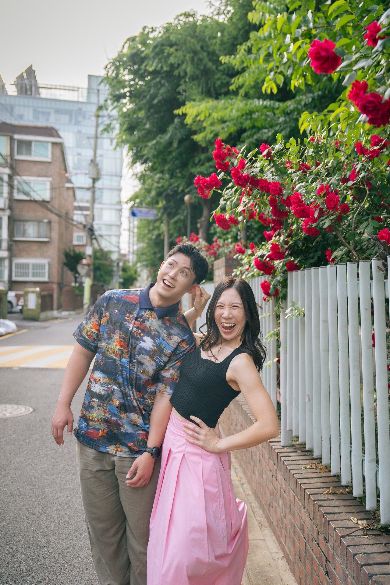 Happy couple laughing together on a street beside a white fence with blooming red roses and lush green trees.