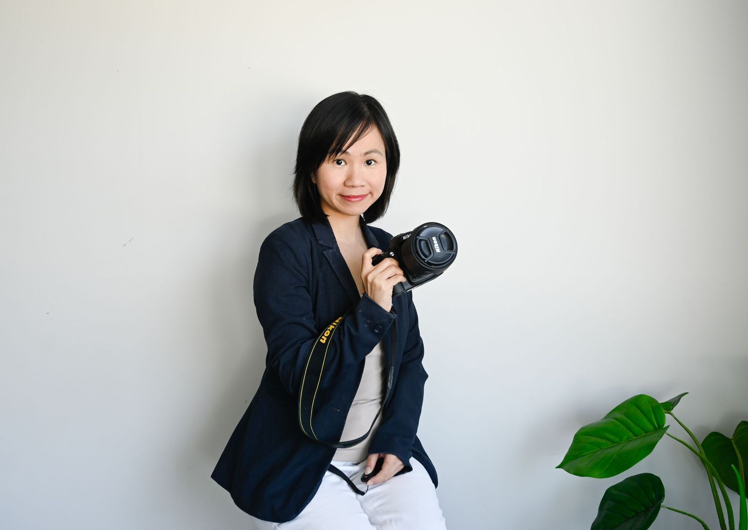 A woman in a beige blazer reviews photos on her DSLR camera while standing against a white wall near a plant.