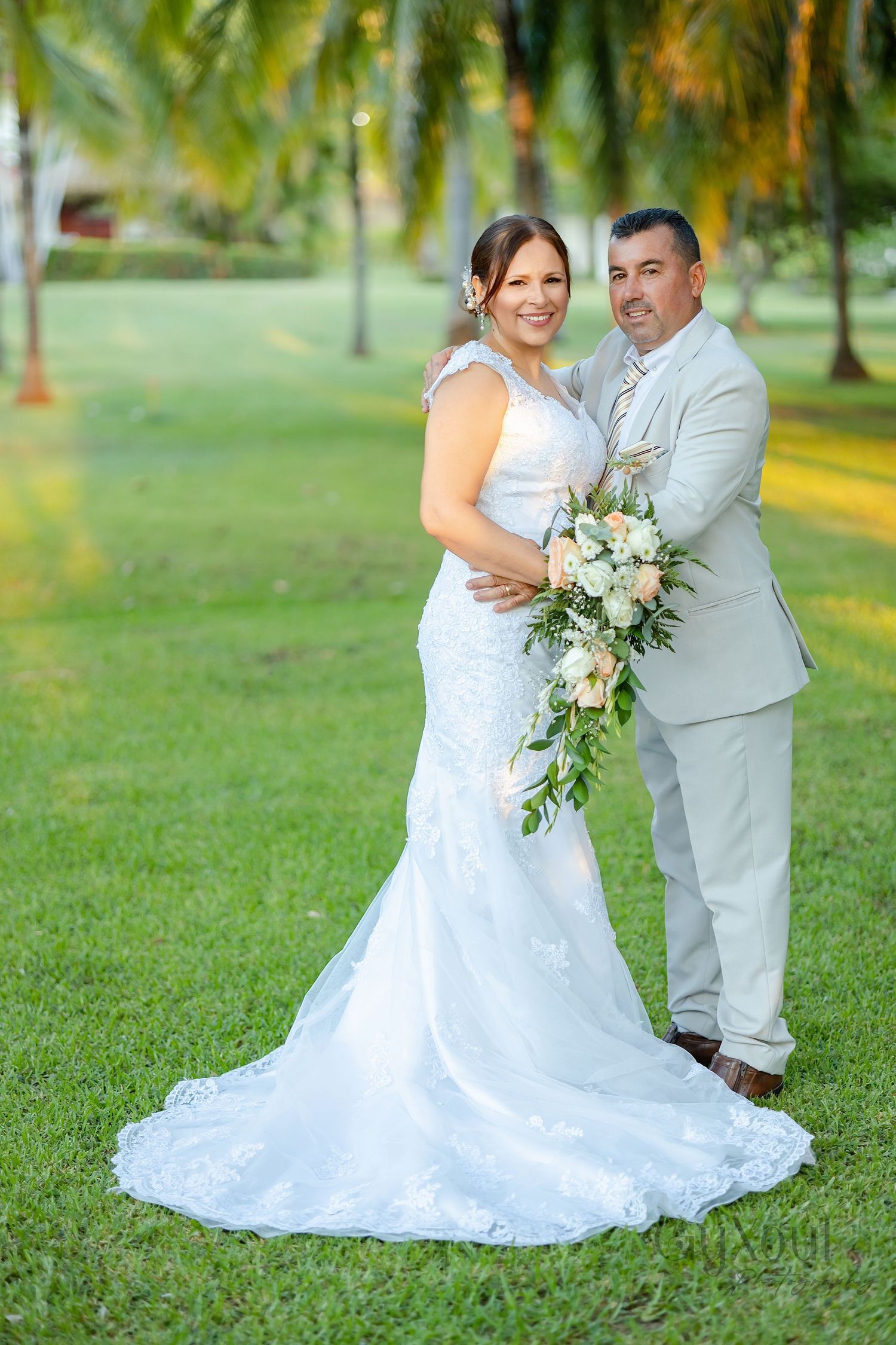 Bride in white lace gown holds bouquet beside groom in tan suit on lush green lawn with palm trees.