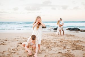 Two children playing on a sandy beach while their parents embrace near ocean waves at sunset.
