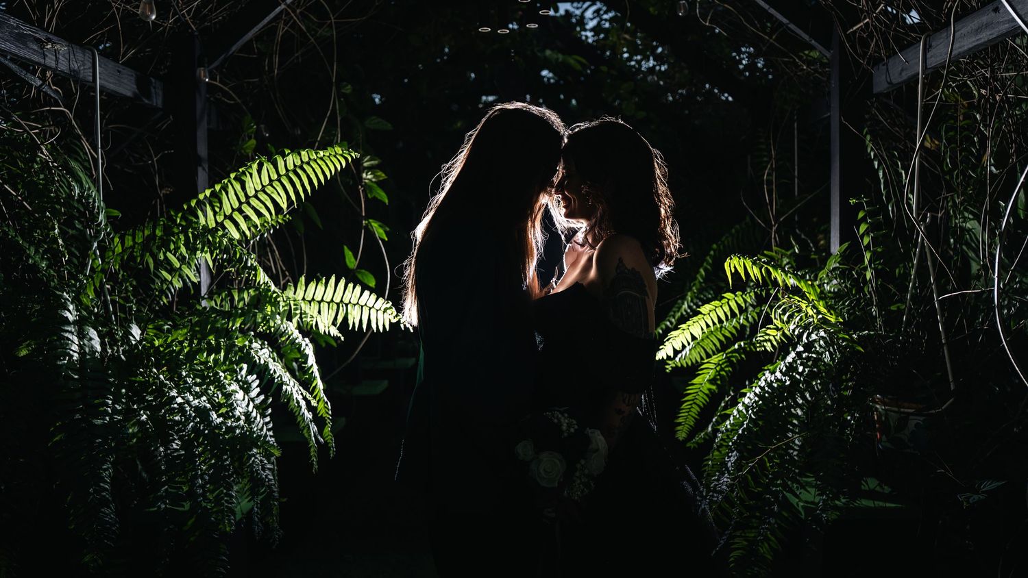 Bride and groom silhouetted among lush green ferns in a dark greenhouse, facing each other intimately.