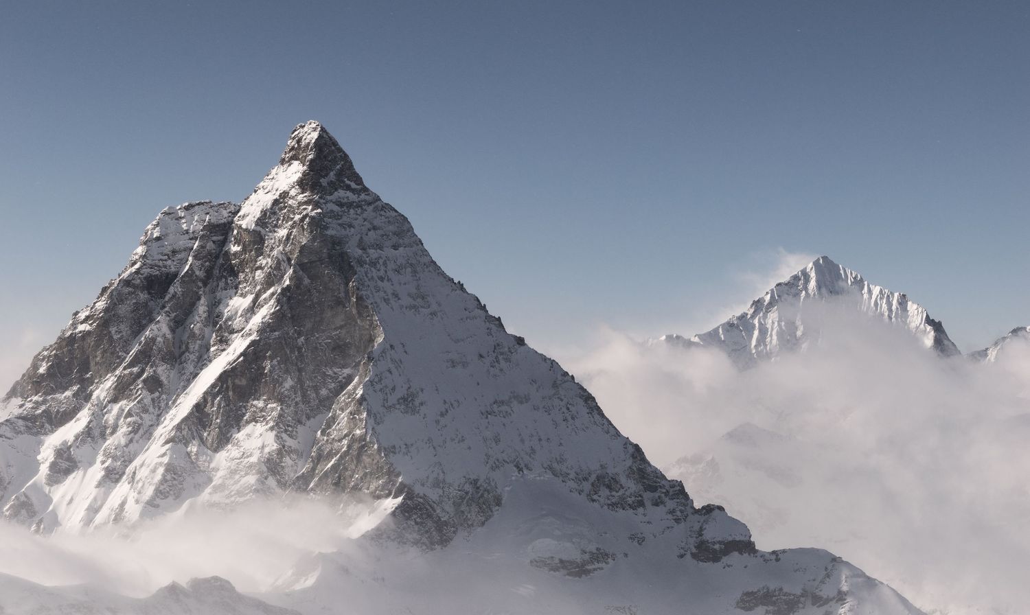Snow-capped mountain peak rising above clouds against a clear blue sky, resembling the Matterhorn in the Alps.