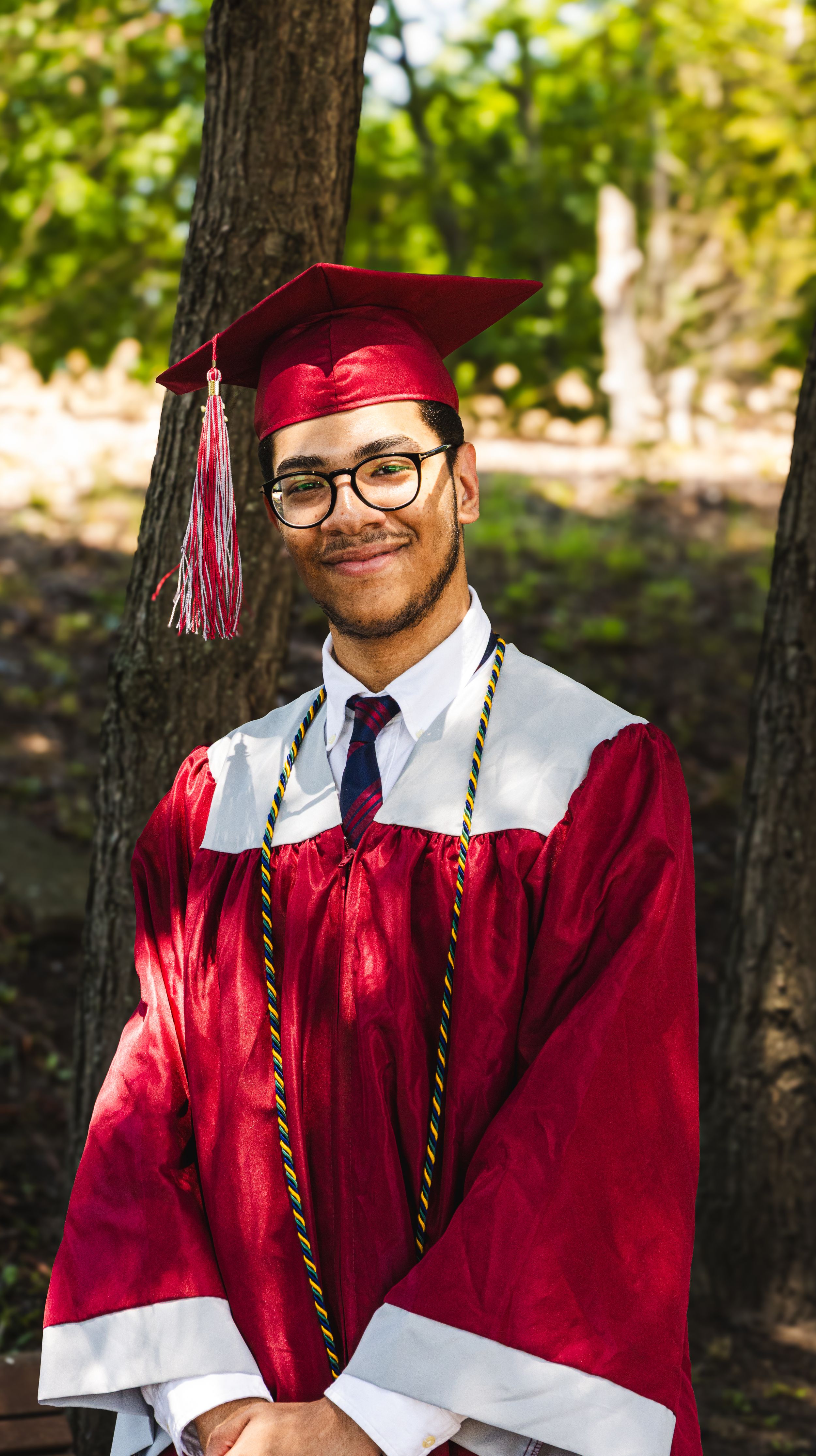 Young man smiling in red graduation cap and gown with honor cords, standing outdoors among trees.