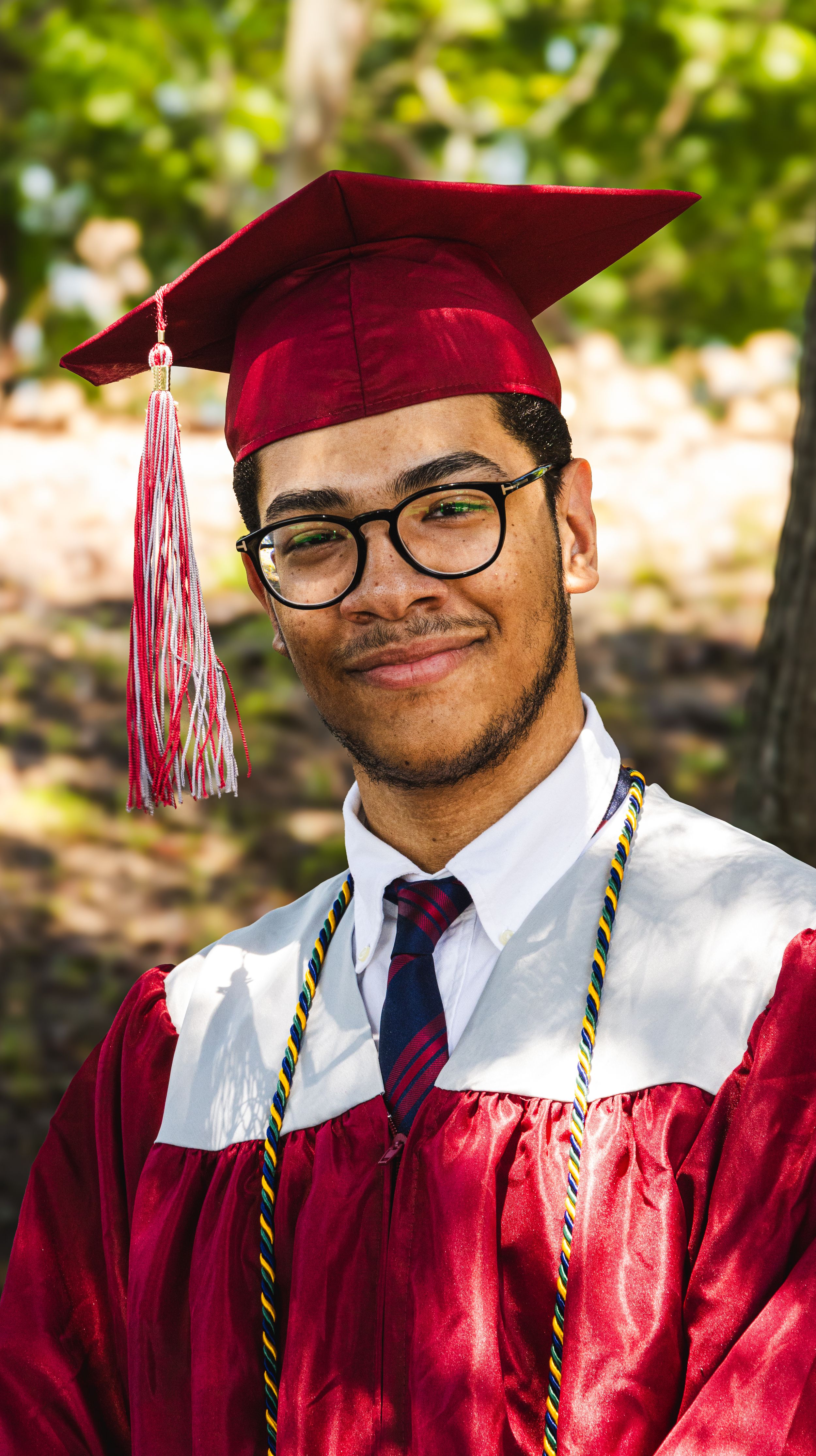 Young man in red graduation cap and gown with glasses and honor cords smiles proudly outdoors.