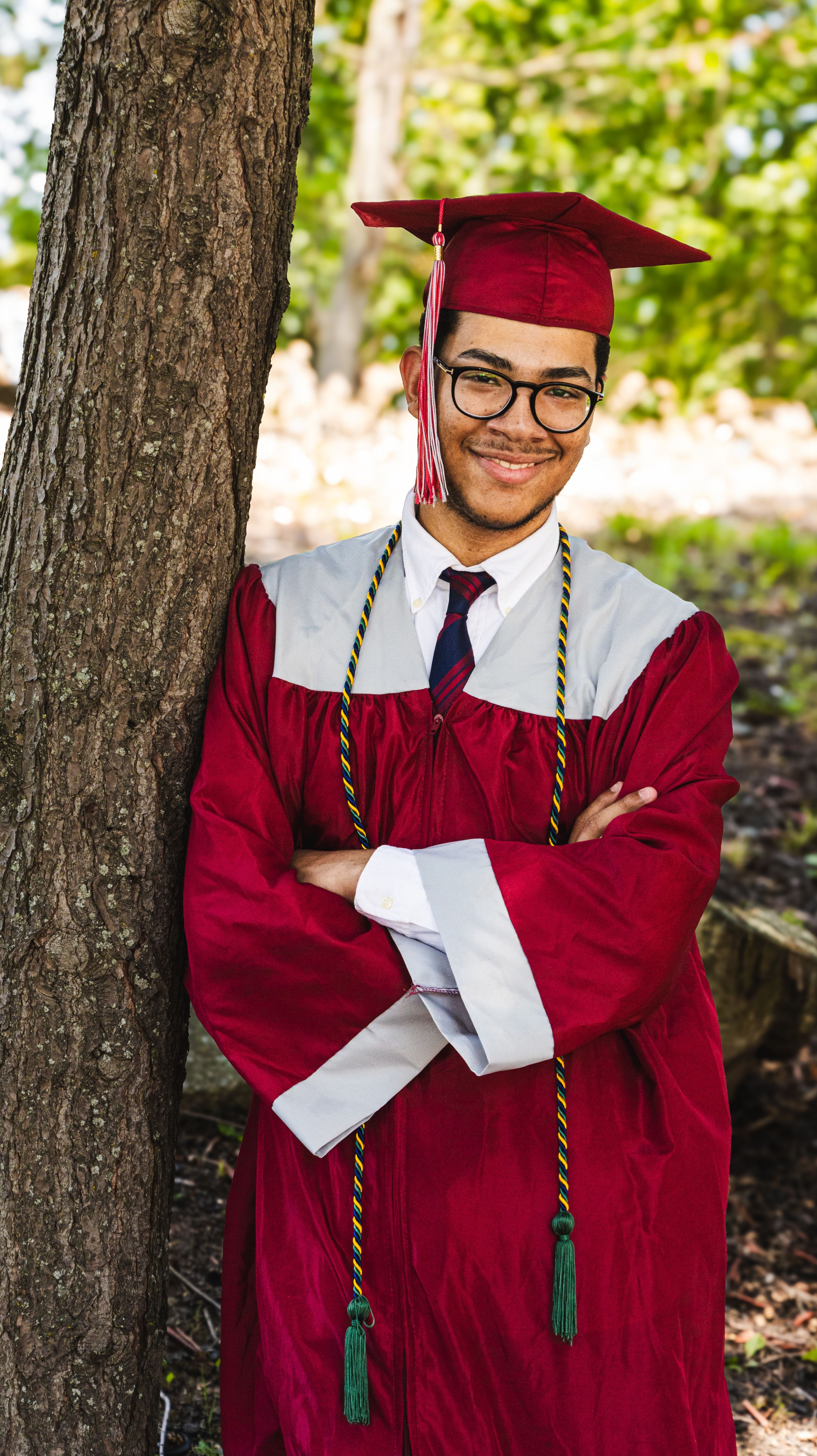 Smiling graduate in red cap and gown with honor cords leans against tree outdoors.