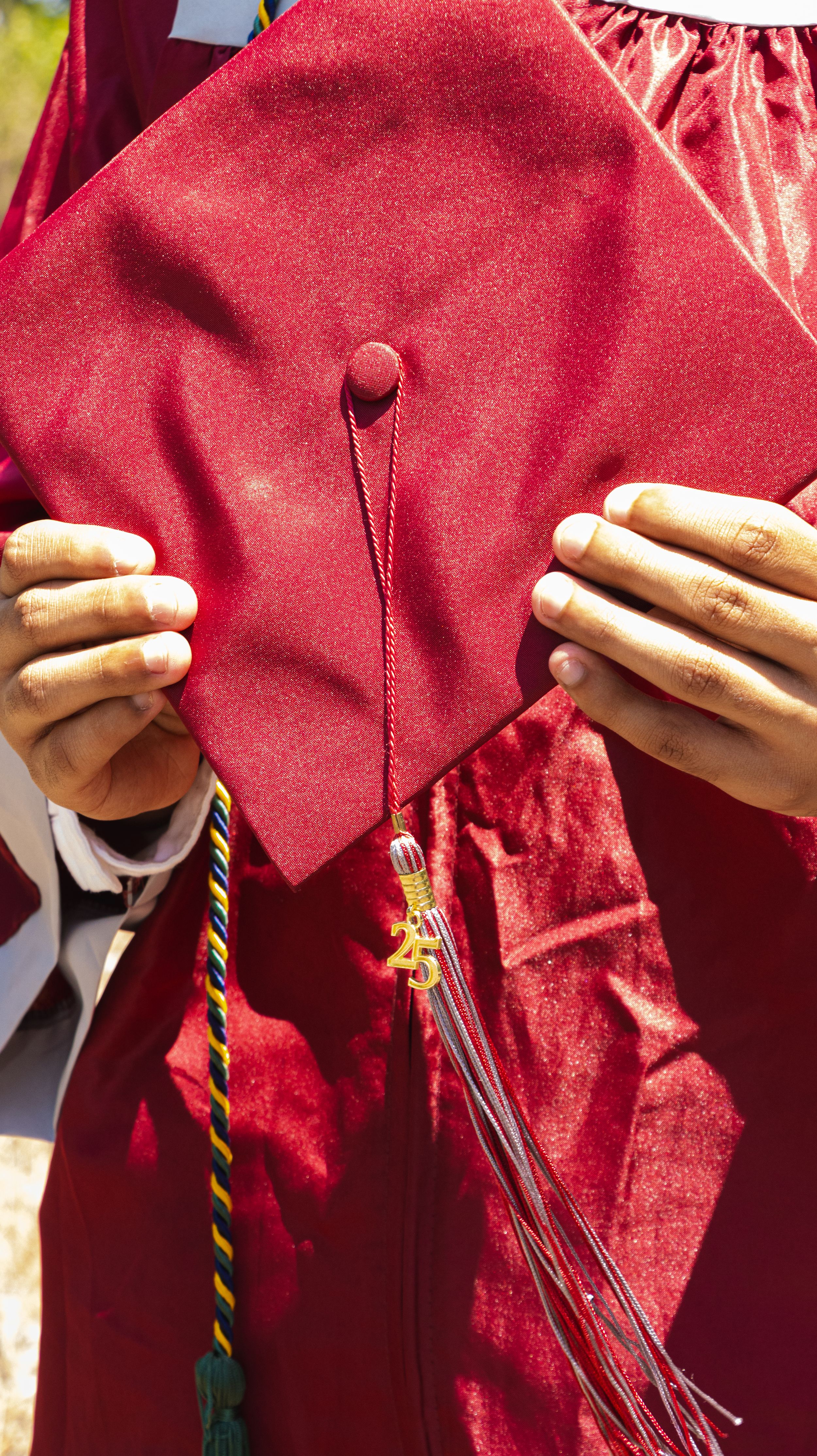 A graduate holds a red graduation cap and gown with colorful honor cords and tassel at an outdoor ceremony.