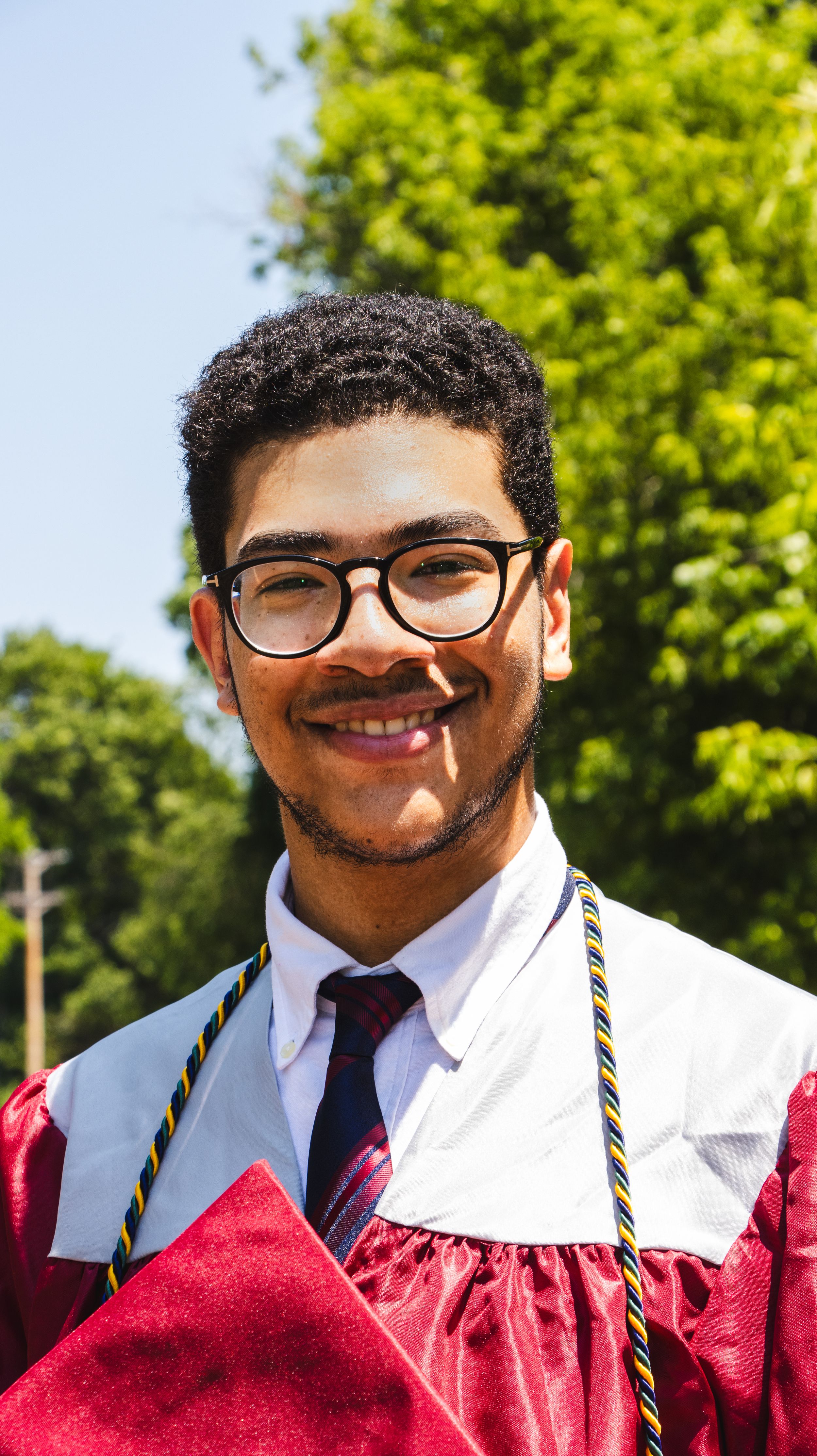 Smiling young man in red graduation gown and glasses poses outdoors with trees in background.