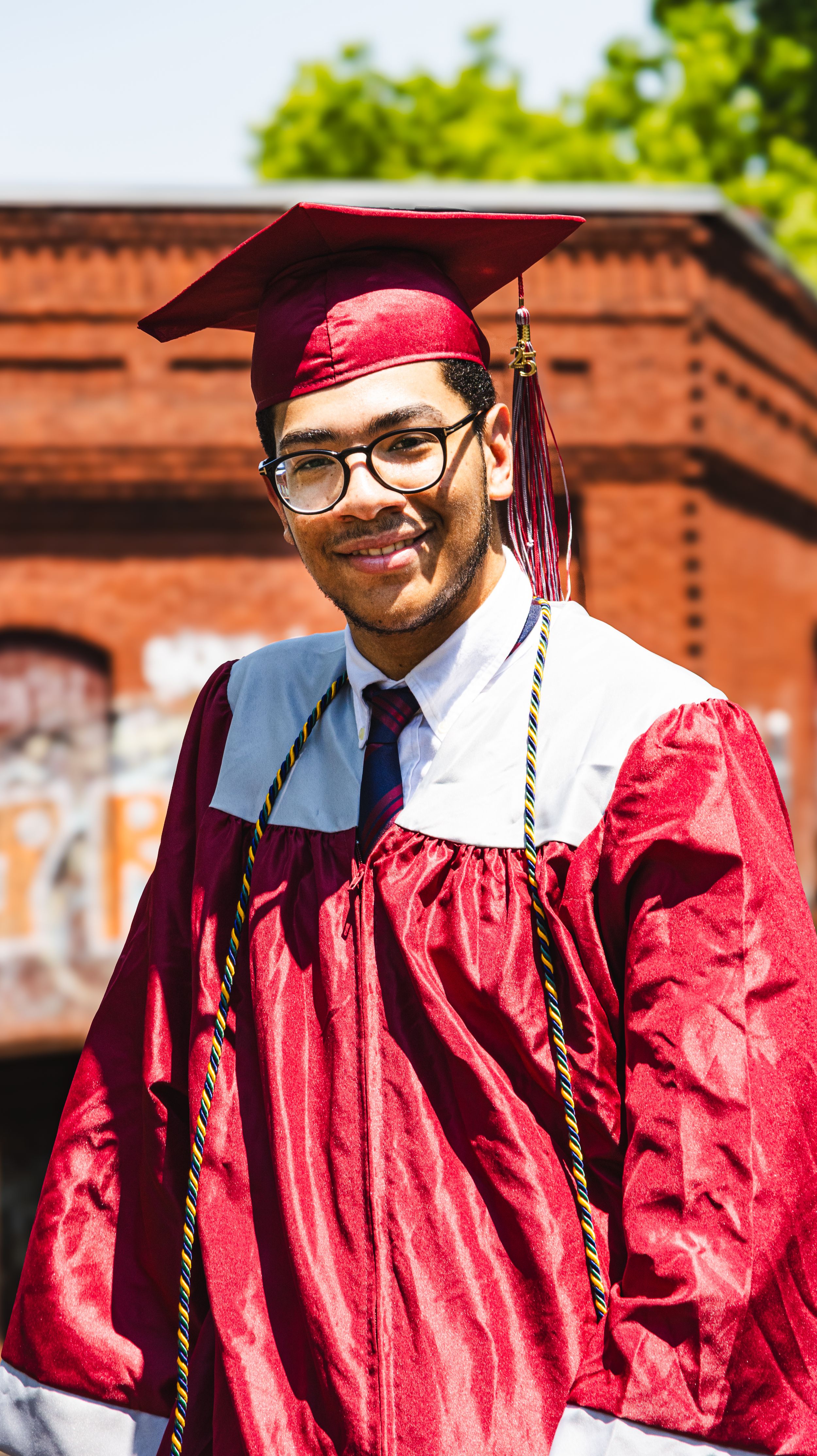 Smiling graduate in red cap and gown with honor cords poses outdoors on a sunny day.