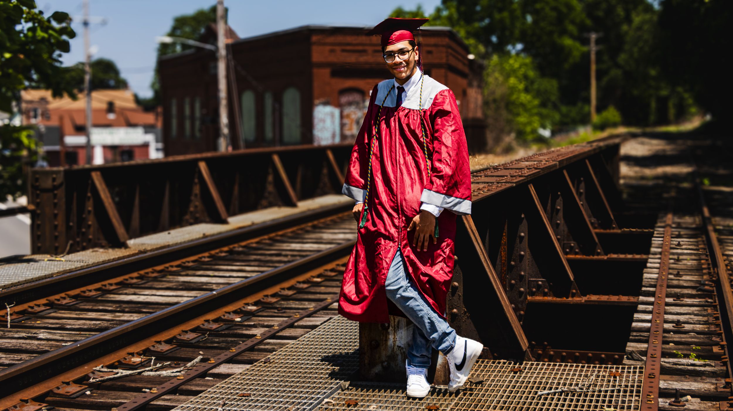 A graduate in red cap and gown poses confidently on railroad tracks in an outdoor senior portrait photo.