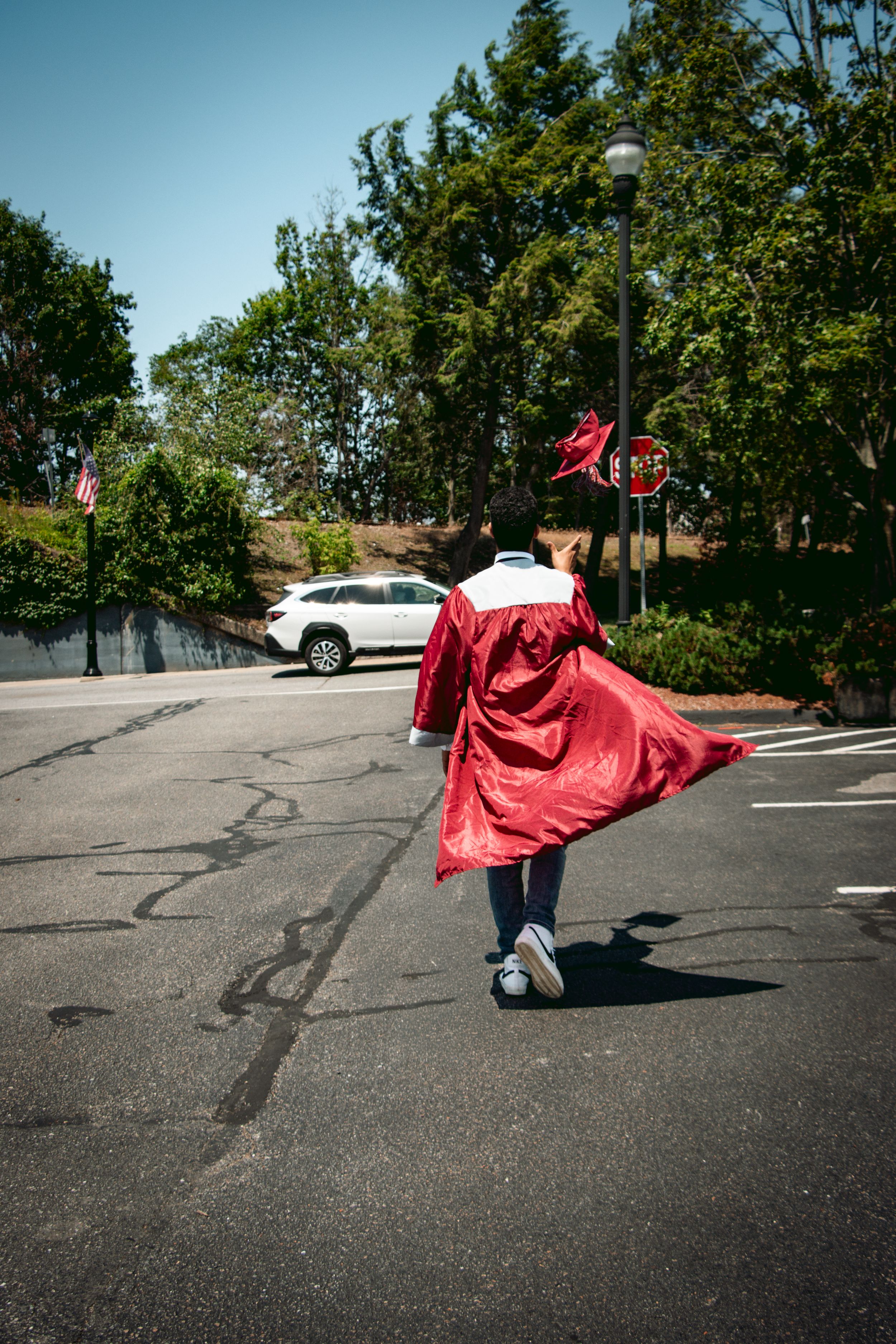 Graduate in red cap and gown walks across a sunny parking lot, with trees and a stop sign in the background.