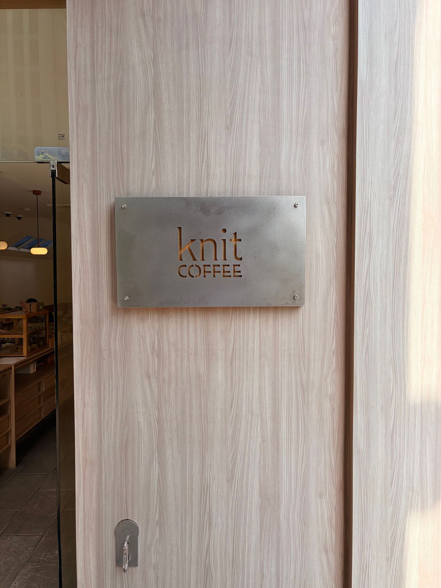 Metal sign reading Knit Coffee mounted on a light wood door entrance to a cozy cafe interior.