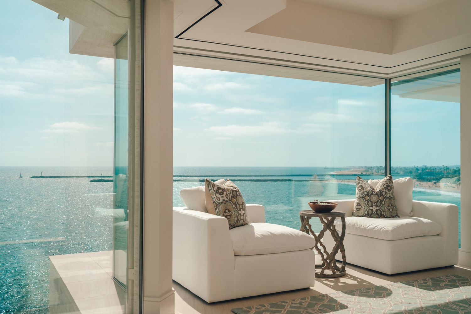 Luxury oceanfront balcony with white armchairs, glass walls, and stunning turquoise sea views on a sunny day.