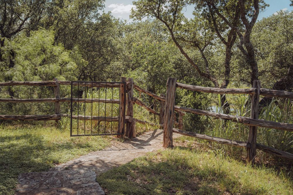 Rustic wooden gate and fence along a stone path surrounded by lush green trees in a natural outdoor setting.