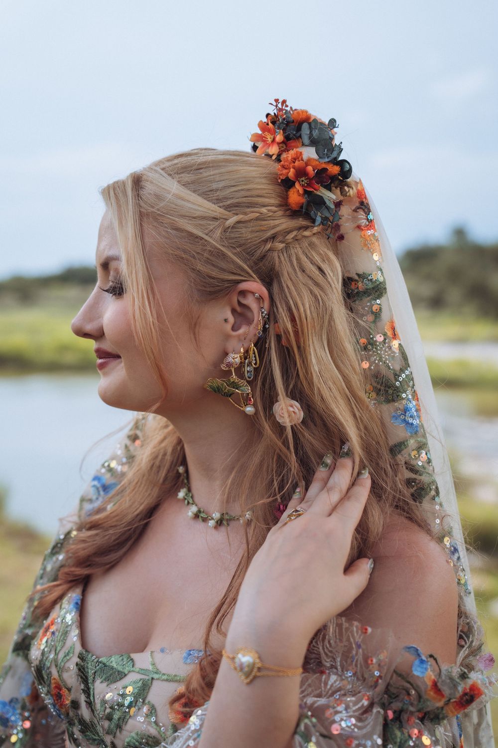 Bride with floral hair accessories, layered earrings, and embroidered veil posing outdoors near water.