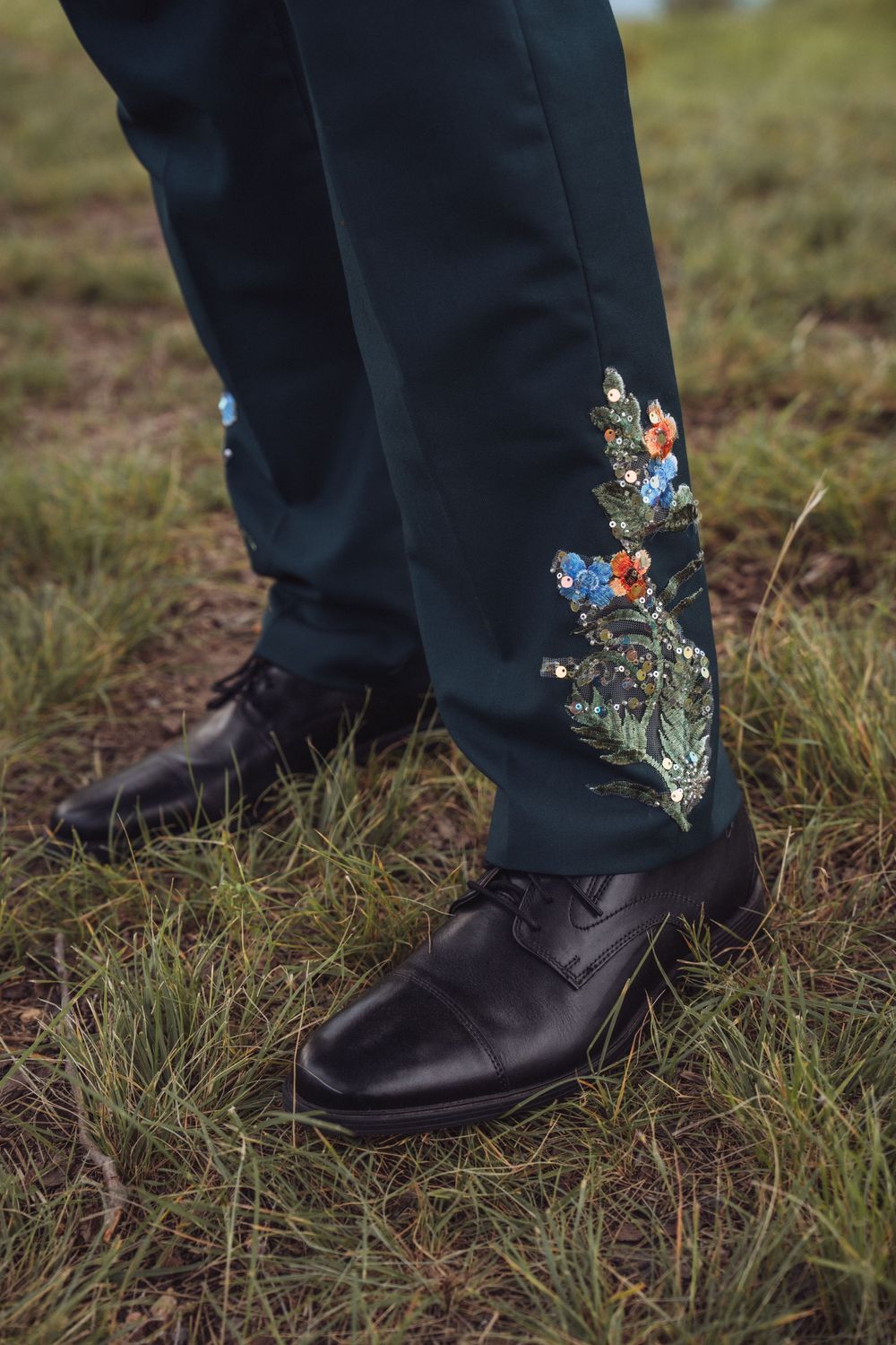 Dark teal embroidered wedding trousers with floral details paired with black leather dress shoes on grass.