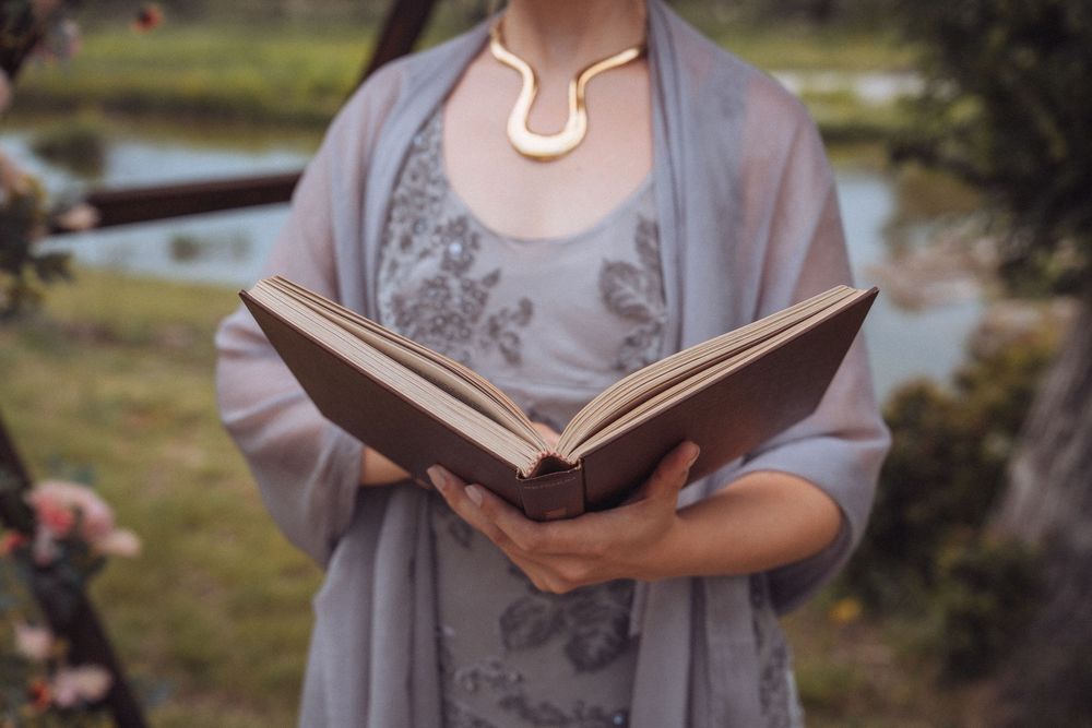 Person in grey shawl holding open brown leather book with gold horseshoe necklace outdoors.