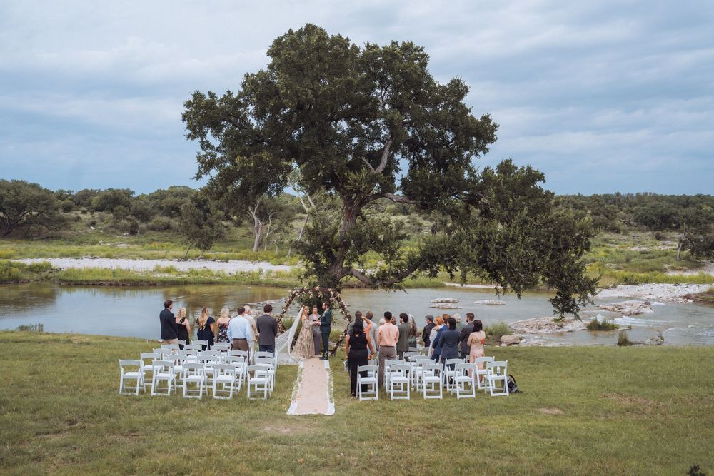 Outdoor wedding ceremony with white chairs arranged near a river under a large oak tree in a scenic landscape.