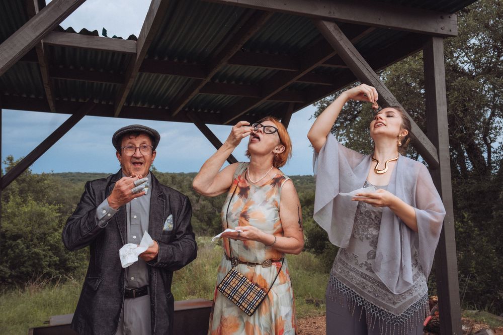 Three elegantly dressed people enjoying drinks outdoors under a rustic wooden pavilion surrounded by lush greenery.