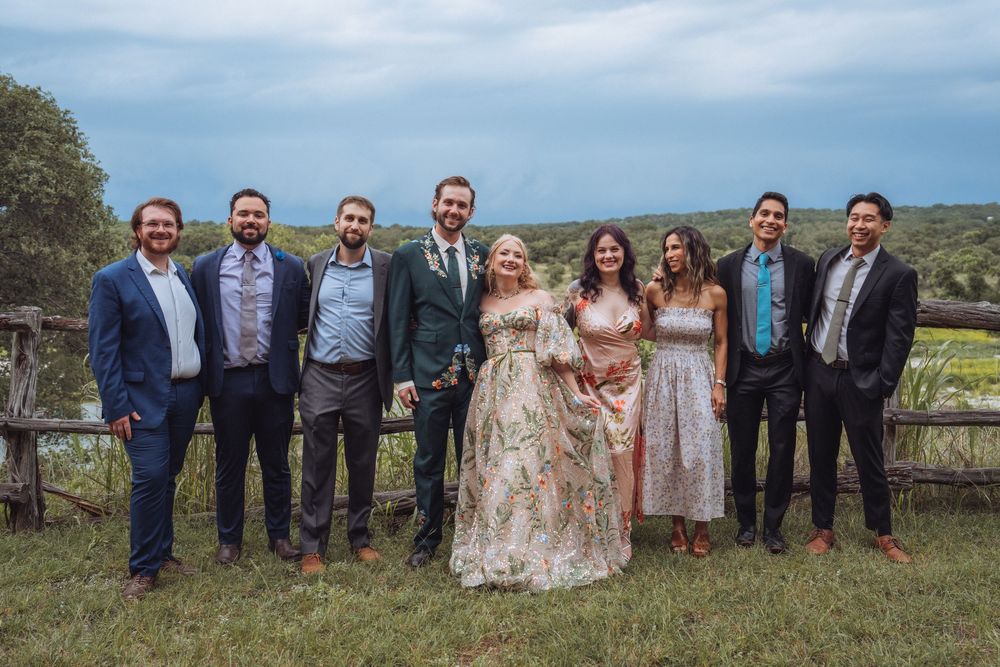 Wedding party of eight people posing together outdoors with a rustic fence and green hills in the background.