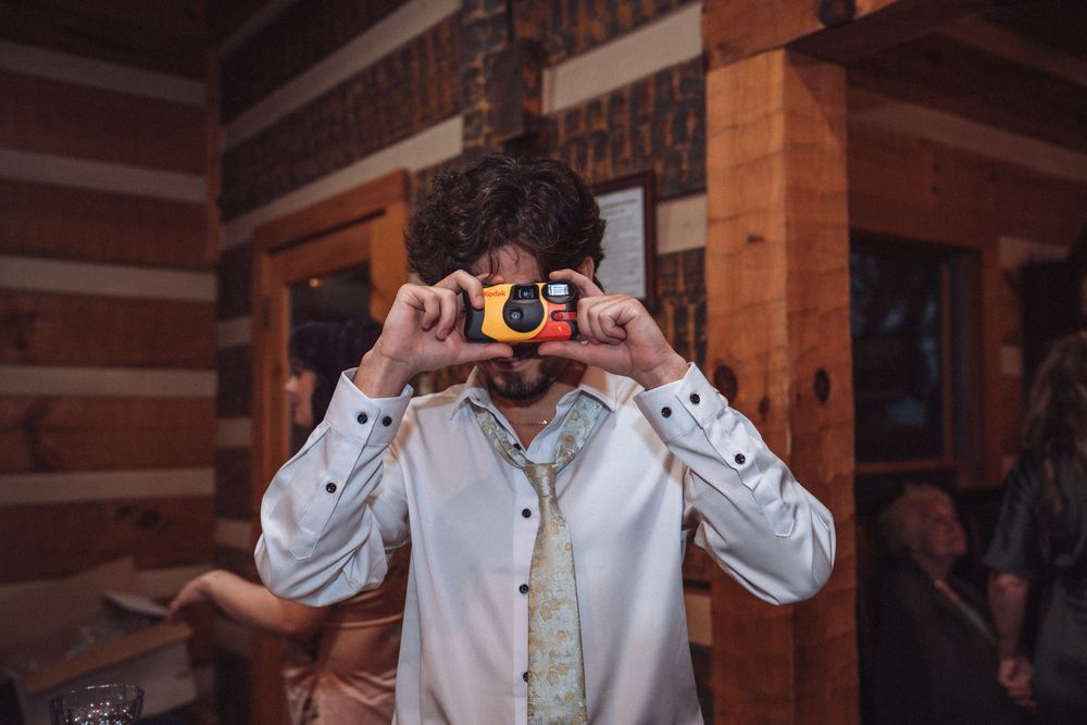 Man in white shirt photographing with yellow disposable camera inside rustic log cabin interior.