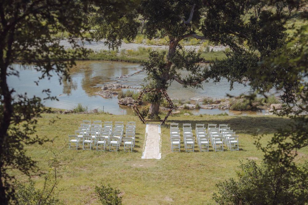 Outdoor wedding ceremony setup with white chairs on green lawn beside a serene river, framed by lush oak trees.