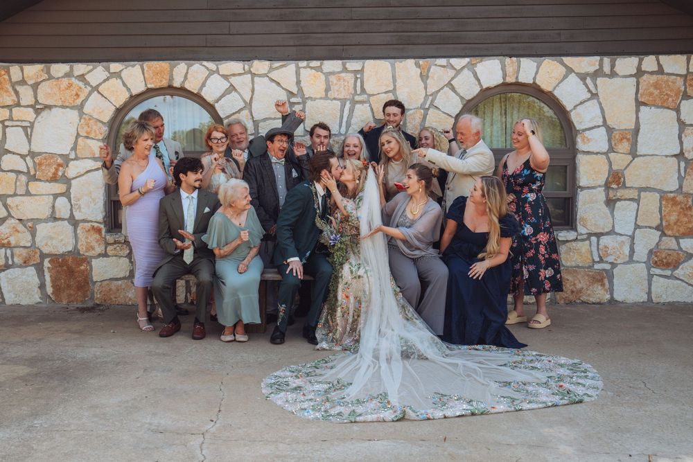 A joyful wedding party poses together in front of a stone wall, with the bride in a lace gown at the center.