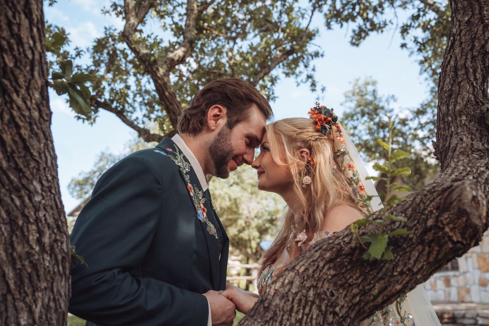 A romantic couple shares an intimate moment touching foreheads under a tree, bride wearing floral hair accessories.