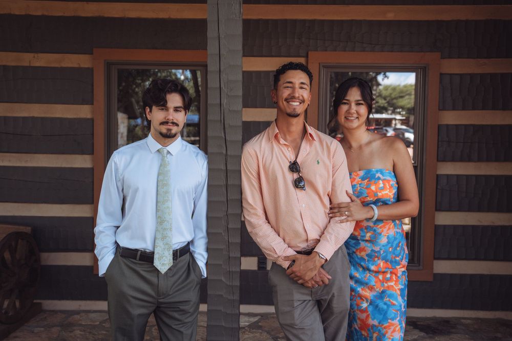 Three people posing outdoors near a wooden building, two men and a woman in colorful floral dress smiling.