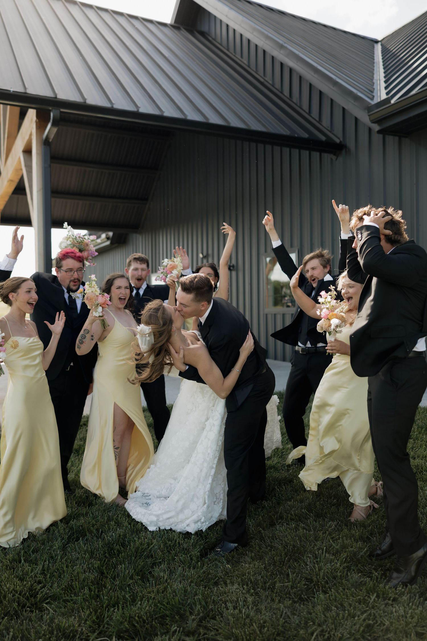 Excited wedding party surrounds bride and groom sharing a kiss outdoors near a gray barn venue.