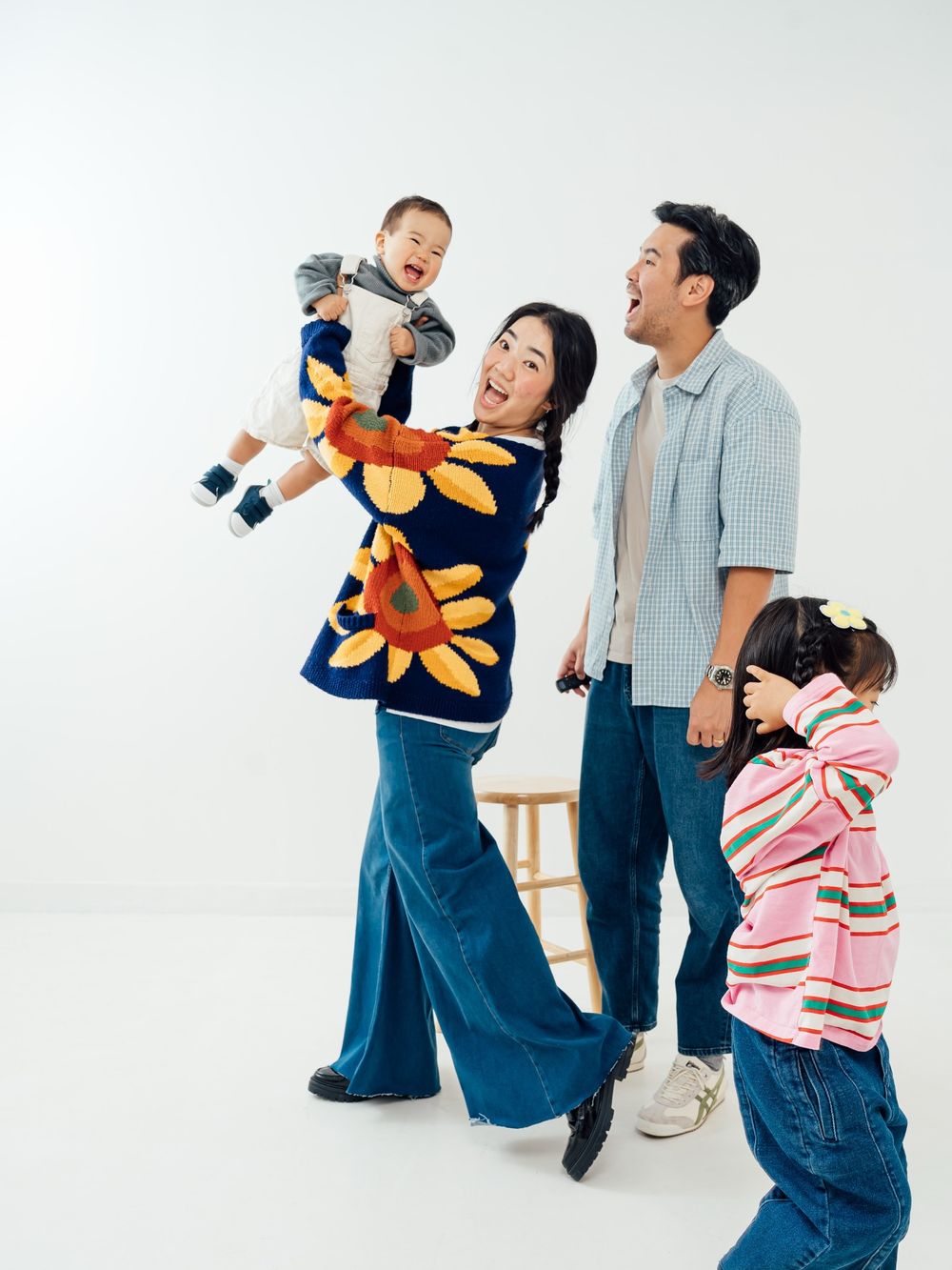 A joyful family of four poses together, with a mom lifting a laughing baby while dad and toddler girl smile on white background.