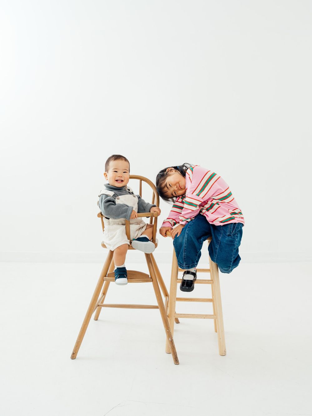 Two young Asian children sit on wooden high chairs against a white background, one leaning playfully toward the other.
